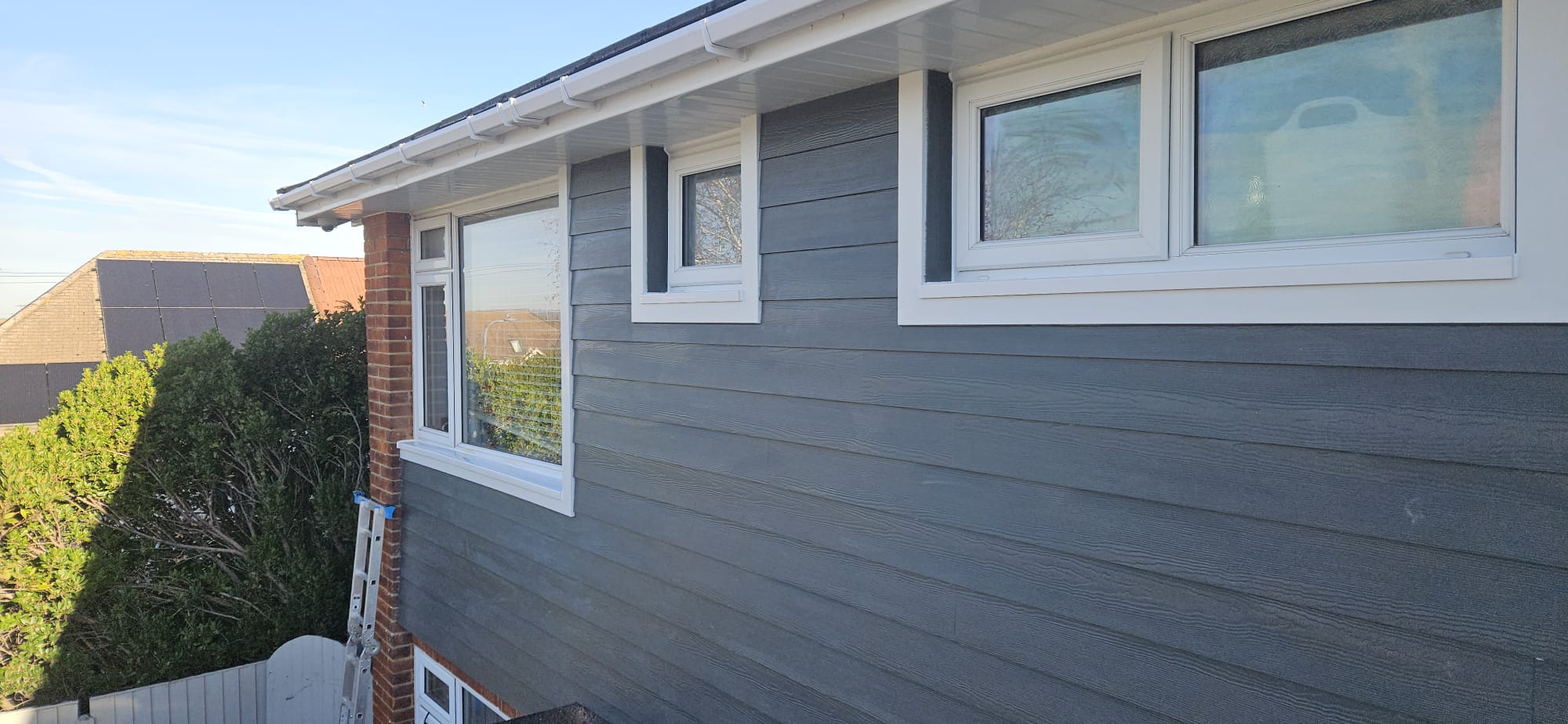 Completed dark grey weatherboard cladding with white window trims, soffit, fascia and guttering on the upper storey of a house